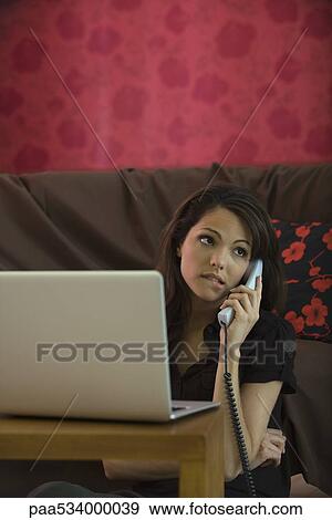Stock Photo - Woman using landline phone and laptop computer in living room, biting lip. Fotosearch