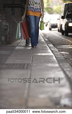 Stock Image - Woman walking on sidewalk, carrying shopping bags, low angle view. Fotosearch