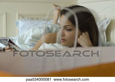 Stock Image - Young woman lying in bed reading magazine, shopping bag in foreground. Fotosearch