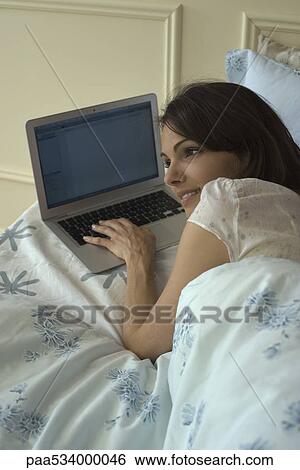 Stock Photograph - Young woman lying in bed, using laptop. Fotosearch