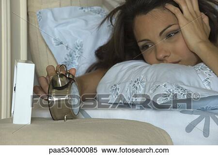 Stock Photography - Young woman silencing alarm clock, holding head. Fotosearch
