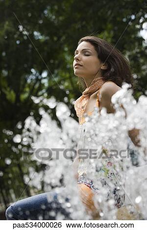Stock Photograph - Young woman sitting by outdoor fountain with eyes closed, low angle view. Fotosearch