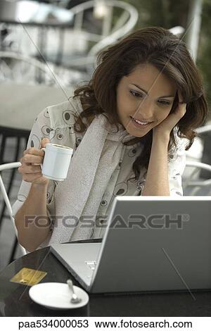 Stock Image - Young woman using laptop in cafe, coffee cup in hand, high angle view. Fotosearch