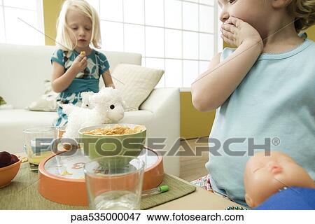 Stock Photo - Little girl looking at bowl of cereal with hand over mouth, sister in background. Fotosearch