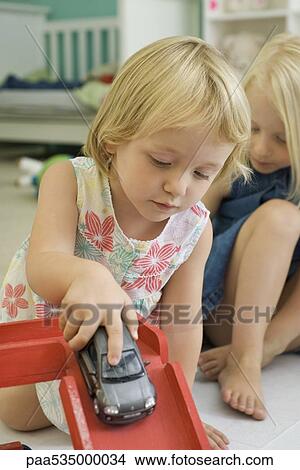 Picture - Little girl playing with toy car, sister in background. Fotosearch