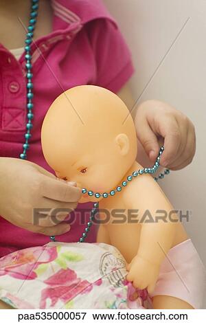 Stock Photo - Little girl putting necklace on baby doll, cropped view. Fotosearch