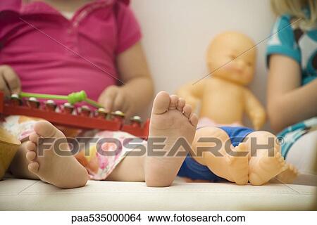 Picture - Little girl sitting beside baby doll, close-up of bare feet. Fotosearch