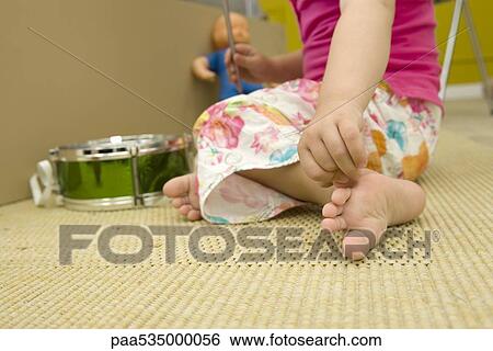 Stock Photograph - Little girl sitting on floor with toys, scratching foot, cropped view. Fotosearch
