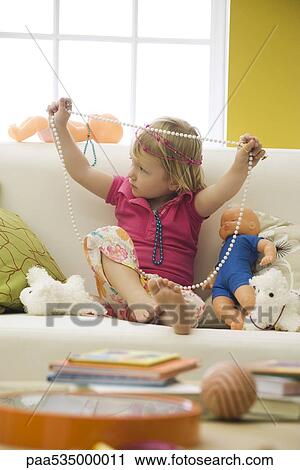 Stock Image - Little girl sitting on sofa with toys, playing with costume jewelry. Fotosearch
