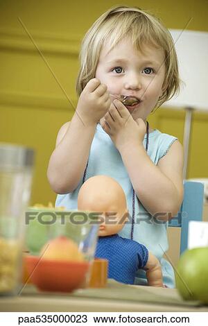 Stock Image - Toddler girl eating at table, baby doll in lap. Fotosearch