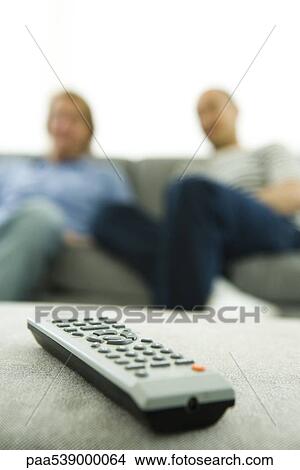 Picture - Close-up of remote control, men sitting on sofa in background. Fotosearch