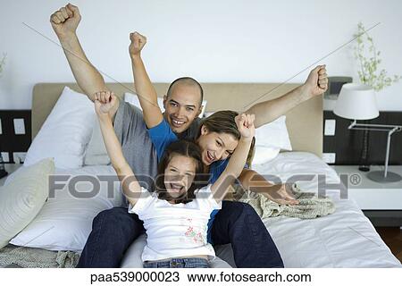 Stock Image - Family sitting together on bed with arms raised in air, all smiling. Fotosearch