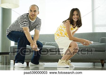 Stock Image - Father and daughter crouching, playing video game with wireless controllers. Fotosearch