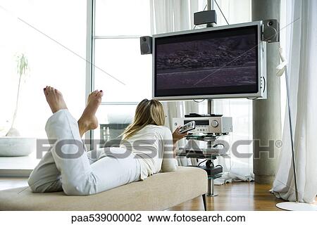 Stock Image - Female lying on chaise longue, watching flat screen television with surround sound. Fotosearch