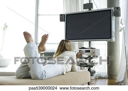 Female lying on stomach watching flat screen TV with surround sound, remote control in hand, rear view View Large Photo Image Picture - Female lying on stomach watching flat screen TV with surround sound, remote control in hand, rear view. Fotosearch