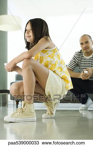 Girl crouching to play video game with wireless controller, father sitting in background View Large Photo Image Stock Image - Girl crouching to play video game with wireless controller, father sitting in background. Fotosearch