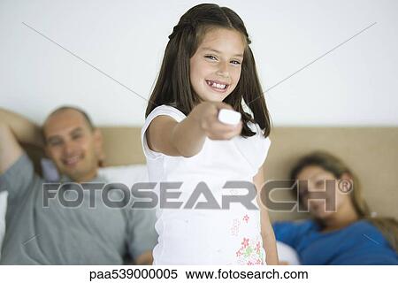 Stock Photography - Girl holding out remote control, smiling at camera, parents in background. Fotosearch