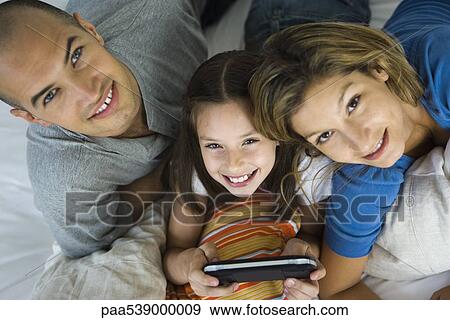 Stock Photo - Girl lying between parents, playing handheld video game, all smiling up at camera. Fotosearch