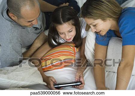Stock Photograph - Girl lying with parents, playing handheld video game. Fotosearch