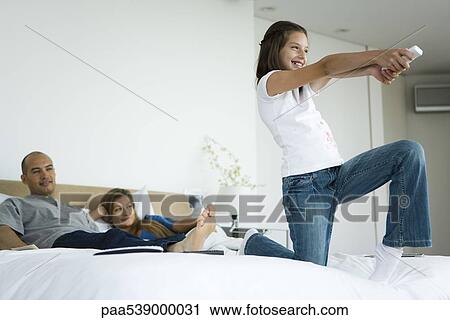 Stock Image - Girl on bed playing video game with wireless controller, parents watching in background. Fotosearch