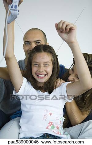 Stock Photograph - Girl sitting with parents, holding video game controller in air and smiling. Fotosearch