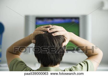 Stock Image - Male watching television, both hands on head, rear view. Fotosearch