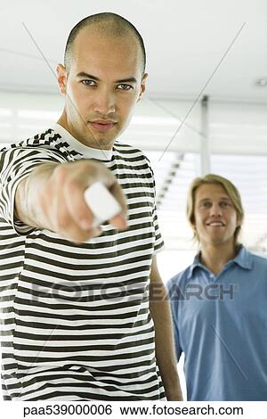 Man pointing remote control at camera, friend standing in background View Large Photo Image Stock Photograph - Man pointing remote control at camera, friend standing in background. Fotosearch