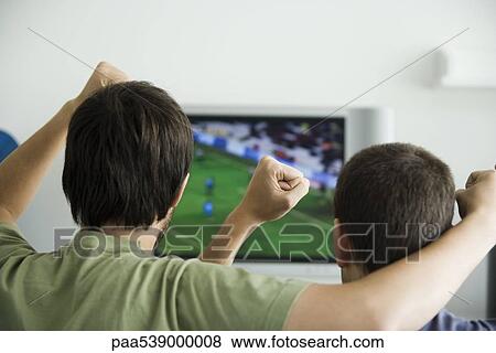 Two males watching sports on television, fists raised in air, rear view View Large Photo Image Stock Photo - Two males watching sports on television, fists raised in air, rear view. Fotosearch