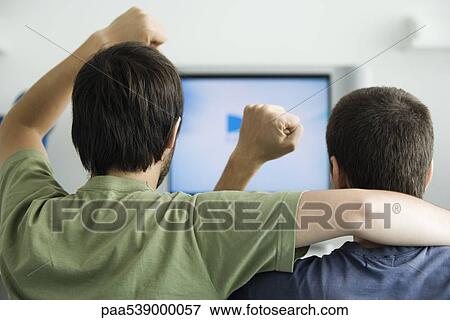 Two males watching TV, fists raised in air, rear view View Large Photo Image Stock Photo - Two males watching TV, fists raised in air, rear view. Fotosearch