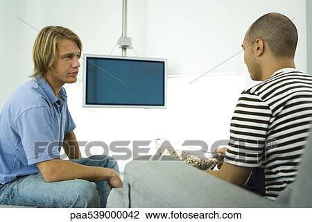 Stock Image - Two men chatting and eating popcorn, flat screen TV in background. Fotosearch