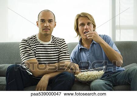 Stock Photo - Two men sitting on sofa eating popcorn. Fotosearch