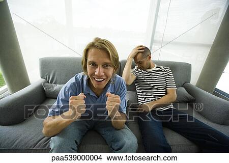 Two men sitting on sofa watching television, one cheering, other holding head View Large Photo Image Picture - Two men sitting on sofa watching television, one cheering, other holding head. Fotosearch