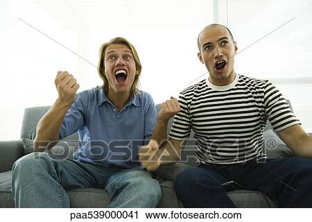Stock Image - Two men sitting on sofa watching TV, cheering with clenched fists. Fotosearch