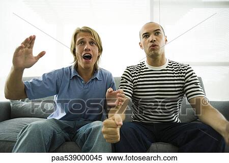 Stock Photography - Two men sitting on sofa watching TV, hands in air. Fotosearch
