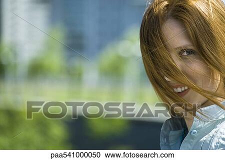 Stock Image - Young red haired woman with hair blowing across face, portrait. Fotosearch