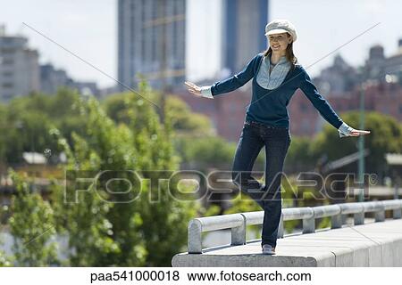 Stock Photo - Young woman in trendy clothing standing on one leg on ledge. Fotosearch