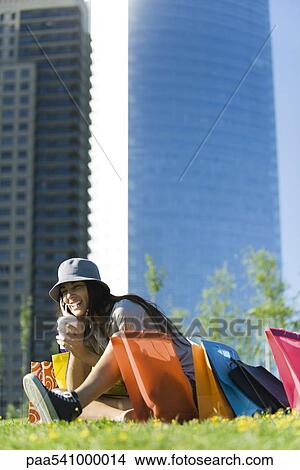 Picture - Young woman sitting in park with shopping bags, sipping drink with straw. Fotosearch