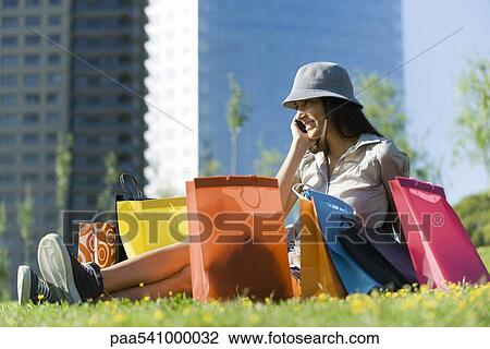 Stock Image - Young woman sitting on ground surrounded by shopping bags, talking on cell phone. Fotosearch