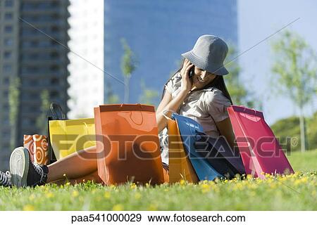 Stock Photo - Young woman sitting on ground surrounded by shopping bags, talking on cell phone. Fotosearch