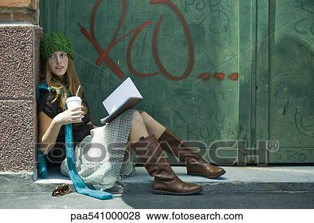 Stock Photo - Young woman sitting on ground with book and drink, looking away. Fotosearch