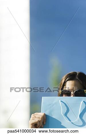 Young woman wearing sunglasses, holding shopping bag in front of face View Large Photo Image Picture - Young woman wearing sunglasses, holding shopping bag in front of face. Fotosearch