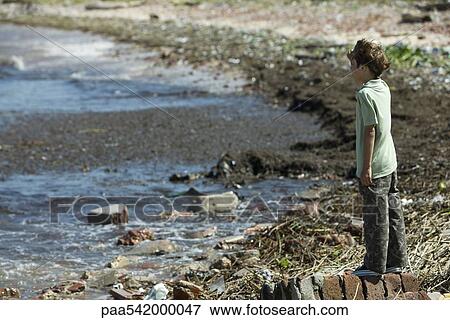 Stock Photo - Boy standing on shore, looking out at polluted water. Fotosearch