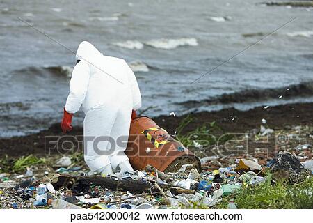 Stock Image - Person in protective suit carrying barrel of hazardous waste on polluted shore. Fotosearch