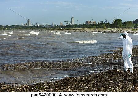 Picture - Person in protective suit standing on polluted shore, city in background. Fotosearch