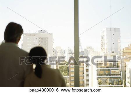 Stock Photograph - Couple, woman's head on man's shoulder, together at window looking at view of city. Fotosearch