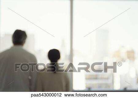 Stock Image - Mature couple looking out window and view of city skyline, rear view. Fotosearch