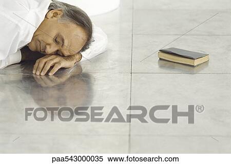 Stock Photography - Senior man lying on tiled floor resting head on arm, sleeping, closed book lying nearby. Fotosearch