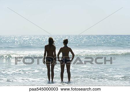 Stock Image - Teen girls standing side by side at the beach, looking at horizon. Fotosearch