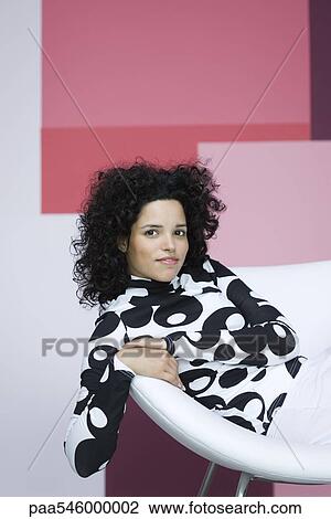 Stock Image - Young woman sitting in chair, smiling at camera, portrait. Fotosearch