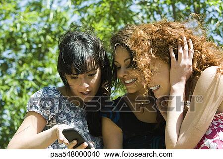 Stock Image - Three young women looking at cell phone outdoors. Fotosearch